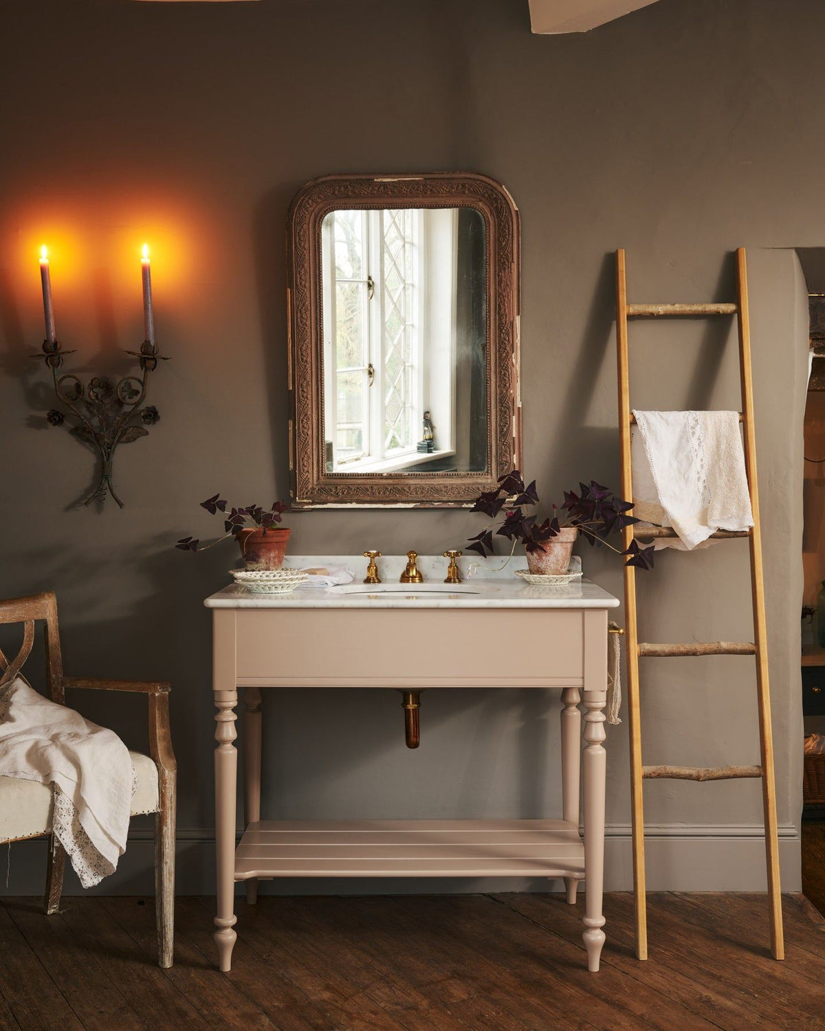 A straight-on view of a dusty-pink Victorian-inspired washstand with a brass mixer tap in a greyish-brown painted room. The shot is atmospheric and moody, with two lit candles casting a subtle glow and light from an adjacent window illuminating the brass accents. | Large Princelet Pink shown TEST