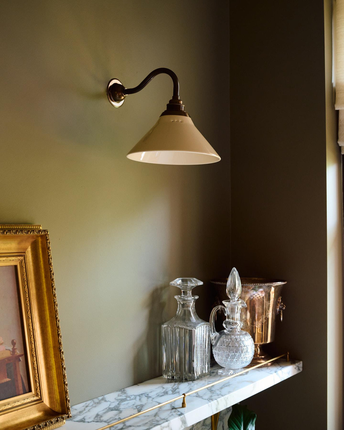 An oblique view of a small wall light with an aged brass swan neck arm and a small cream glazed ceramic shade, fixed to a pink brick wall beside a linen curtain hanging from an aged brass rail. TEST