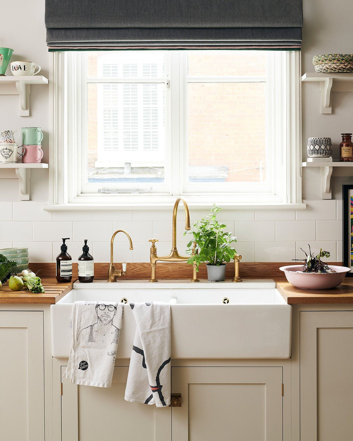 A close-up view of a wide ceramic Belfast sink in a Shaker-style kitchen with creamy mushroom painted cupboards, aged brass taps and a light hardwood worktop. TEST