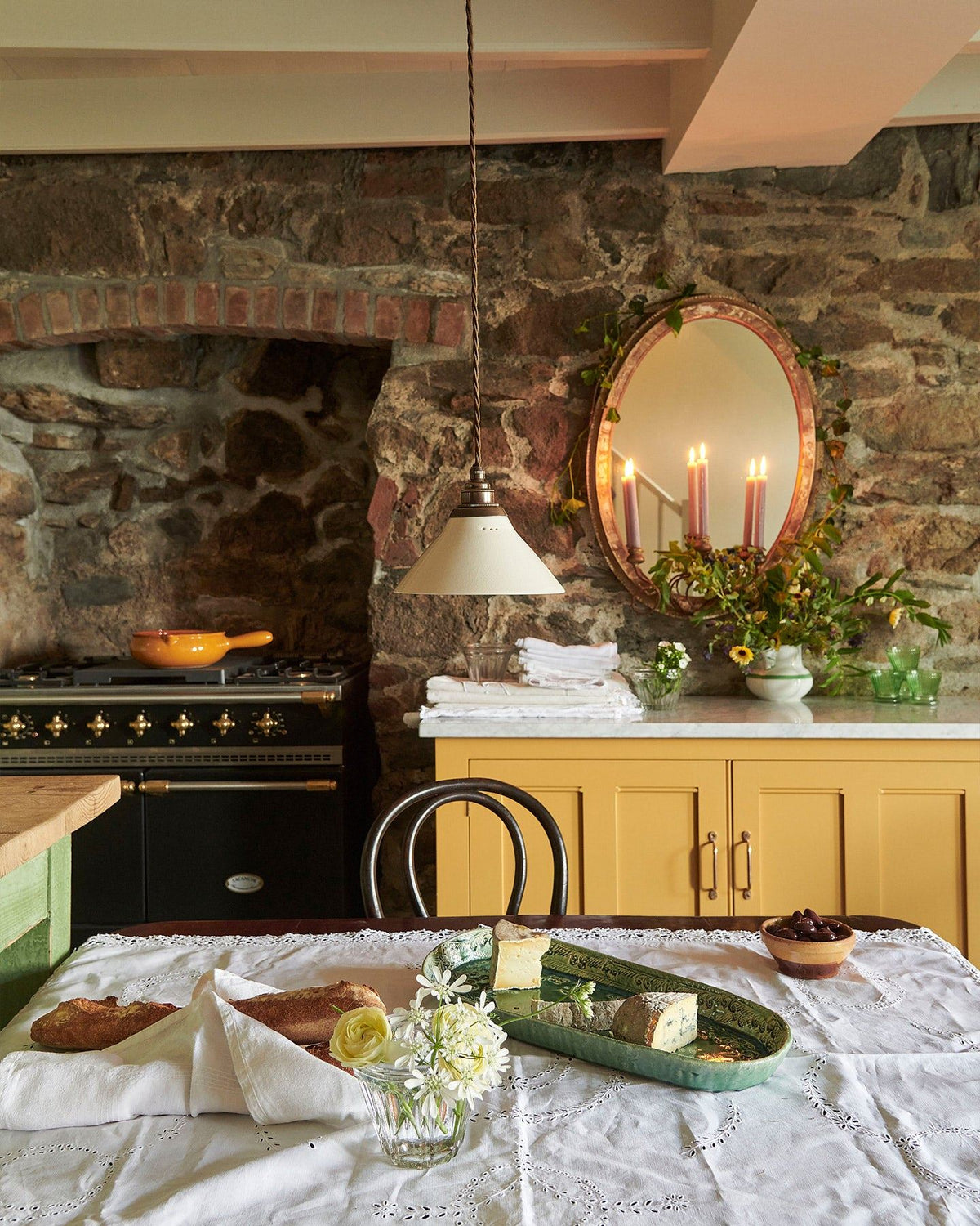 Looking over a table with a vintage lace tablecloth and bohemian ceramic platter at yellow painted Georgian-style cupboards either side of a black range cooker. A stone wall with vintage mirror are seen above. TEST
