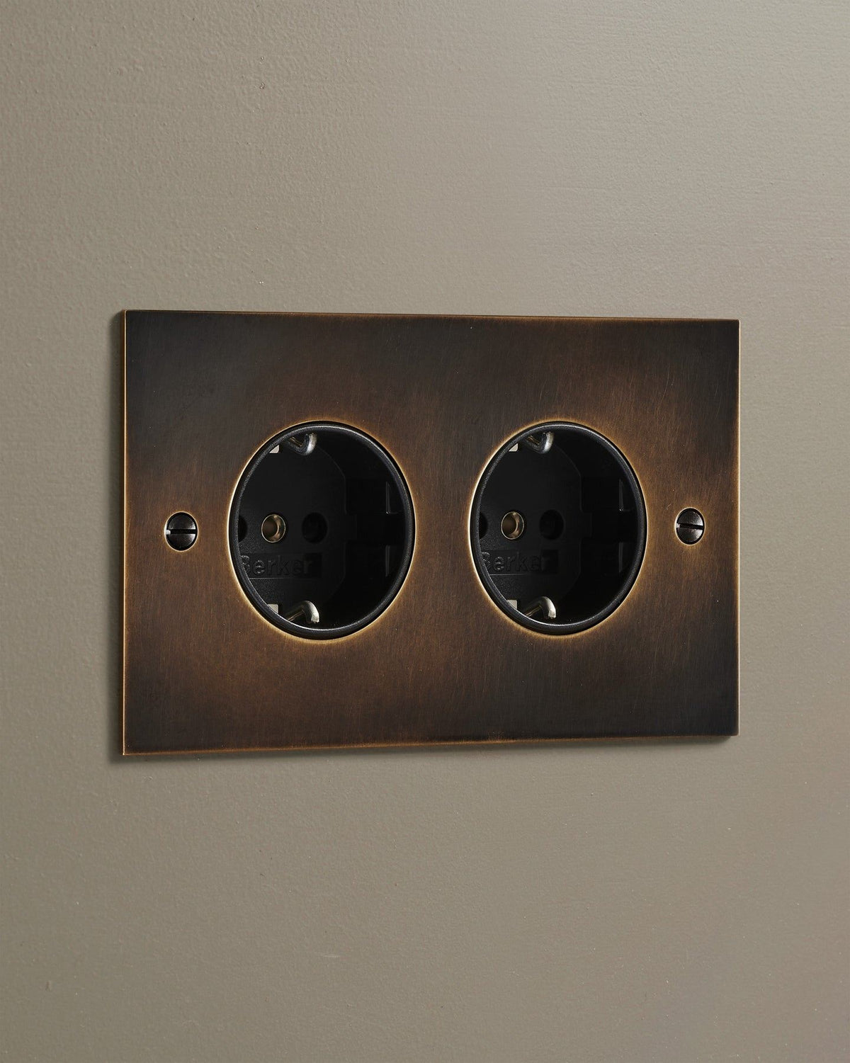 An oblique view of a classic Schuko-style twin plug socket in a dark oxidised brass finish. TEST