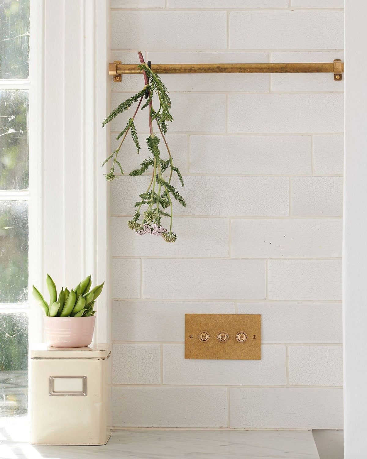 A straight-on view of a 3 gang classic toggle switch in an aged brass finish on white metro tiles with a small olive branch dangling from a short aged brass hanging rail above. | 3 Gang shown TEST