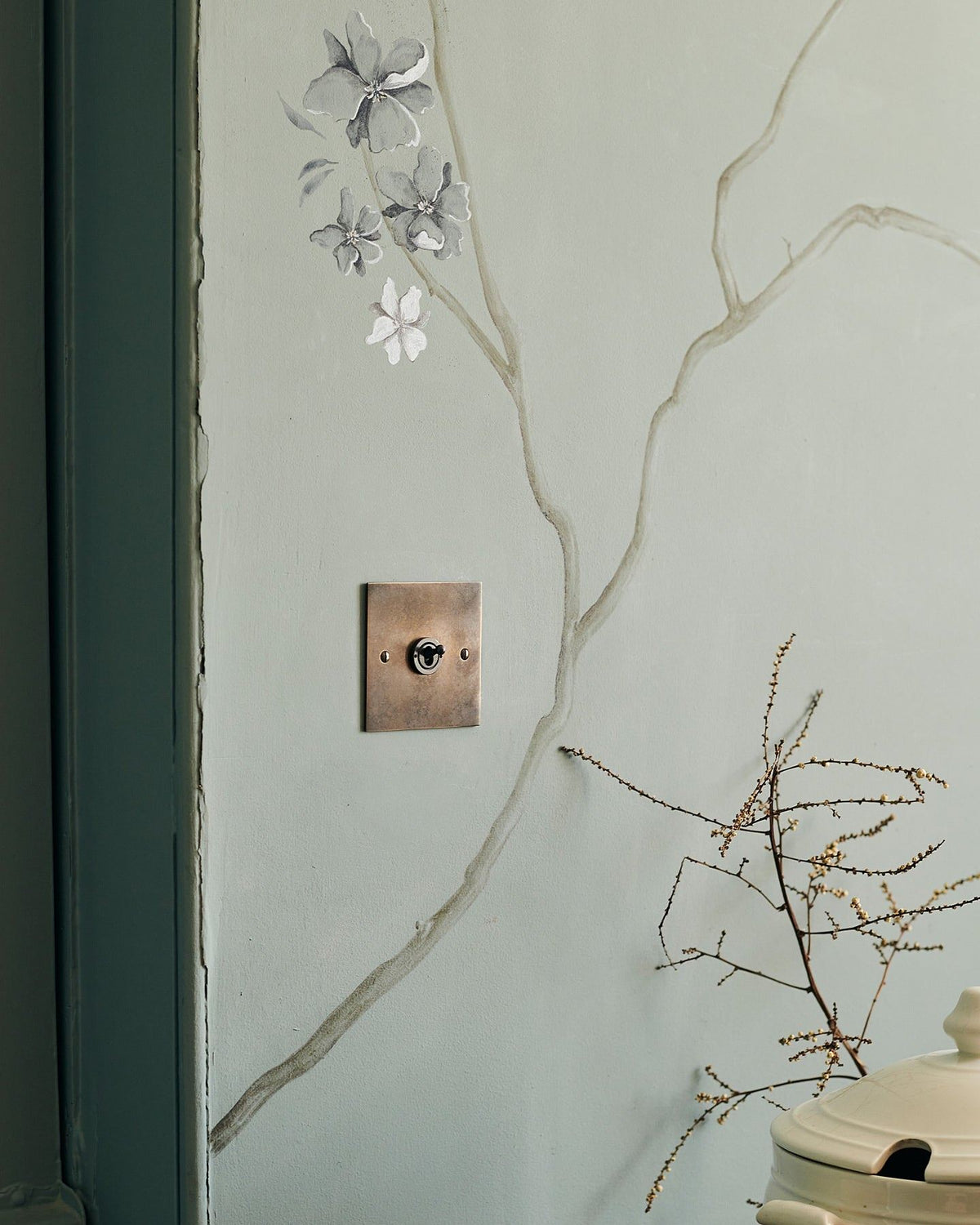 An oblique view of a 1 gang classic toggle switch in a dark oxidised brass finish on a duck-egg blue wall with a delicate handpainted mural of winding branches. | 1 Gang shown TEST
