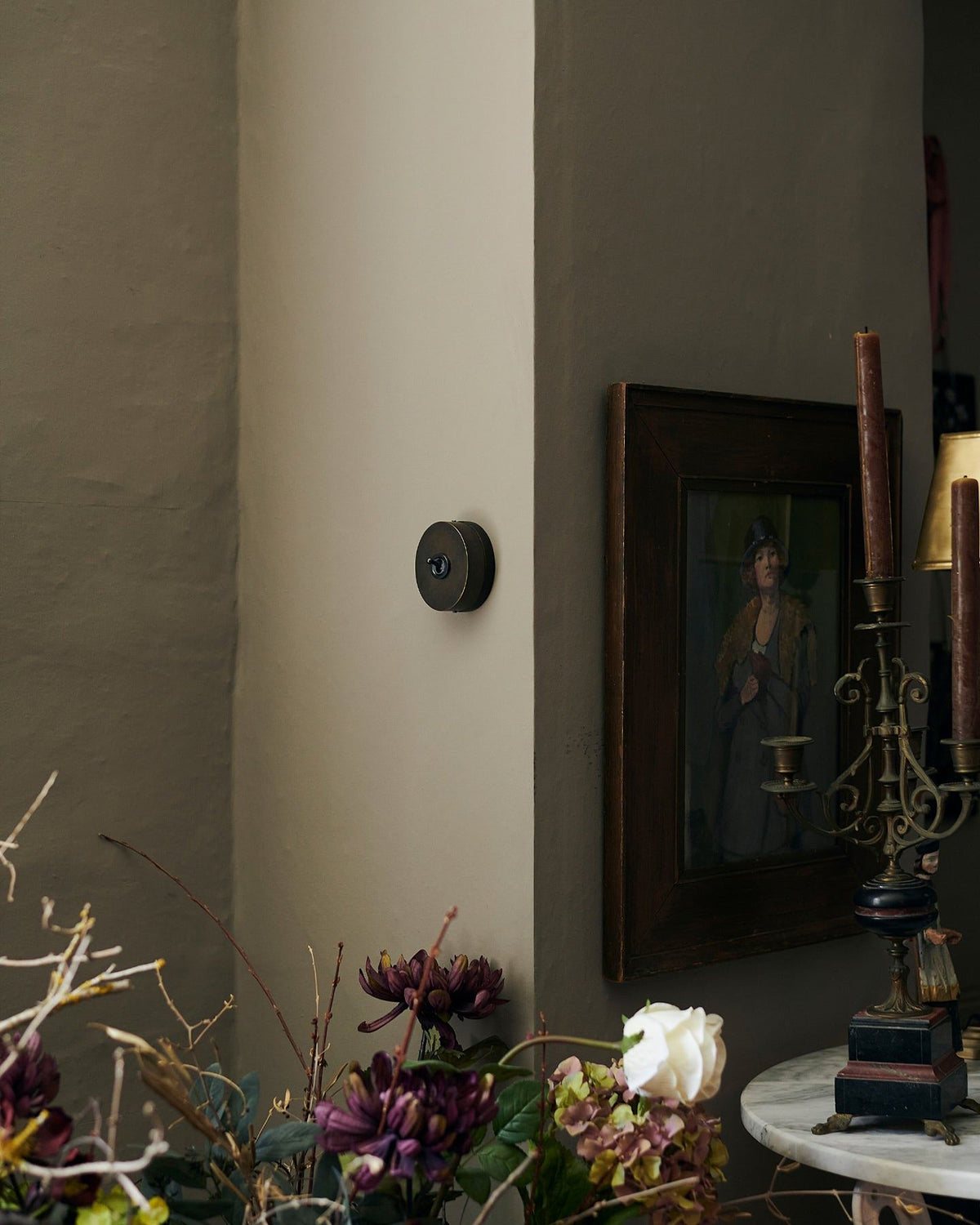 A small 1 gang oval-shaped toggle switch in a dark oxidised brass finish on a grey wall, a moody impressionist oil painted hangs on the wall around a corner with dried flowers in the foreground. TEST