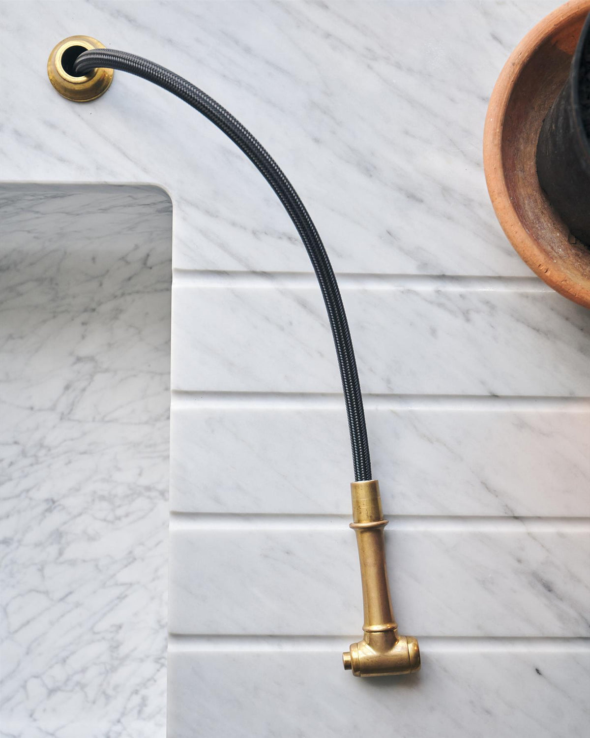 An overhead view of a traditional-style aged brass rinser tap and black hose, pulled out and resting on a Carrara marble worktop. TEST