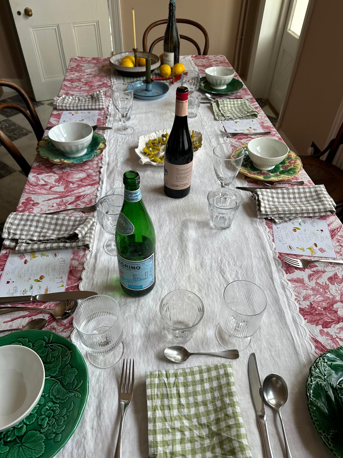 A dining table laid with a tablecloth, a mix of plates, check napkins, wine glasses and a few bottles and plates of food.