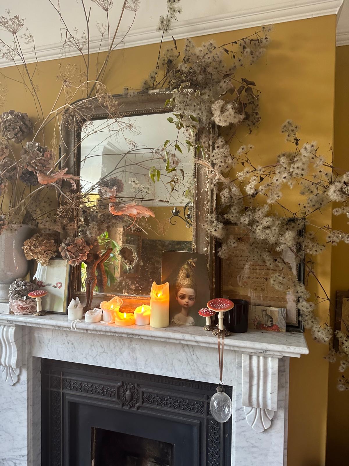 A marble fireplace with lots of little trinkets on the mantelpiece, as well as dried flowers and foliage. A large mirror has been propped against the rich mustard yellow wall behind.