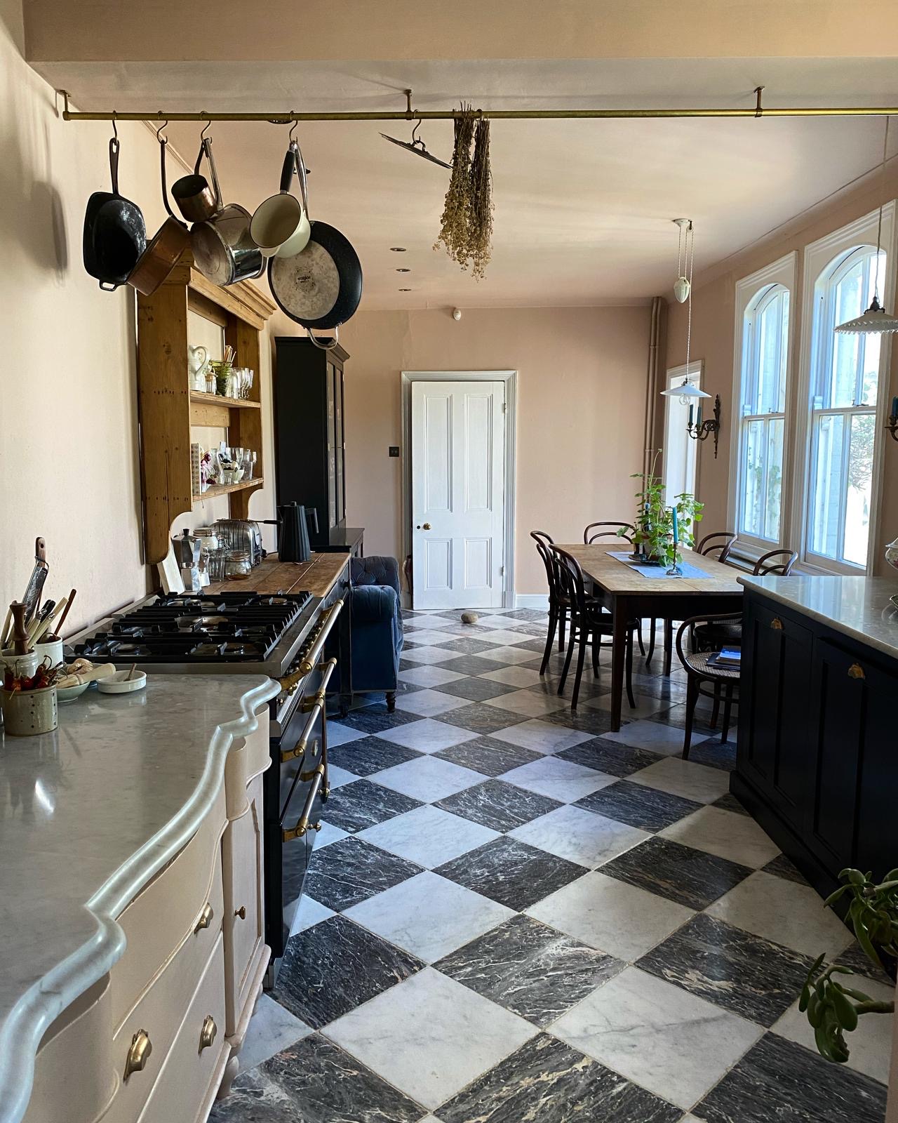 A full room shot of a peaceful looking kitchen. The walls are soft pink and there is a selection of freestanding furniture pieces, as well as a black range cooker, down each wall. Black and white chequerboard tiles make up the floor.