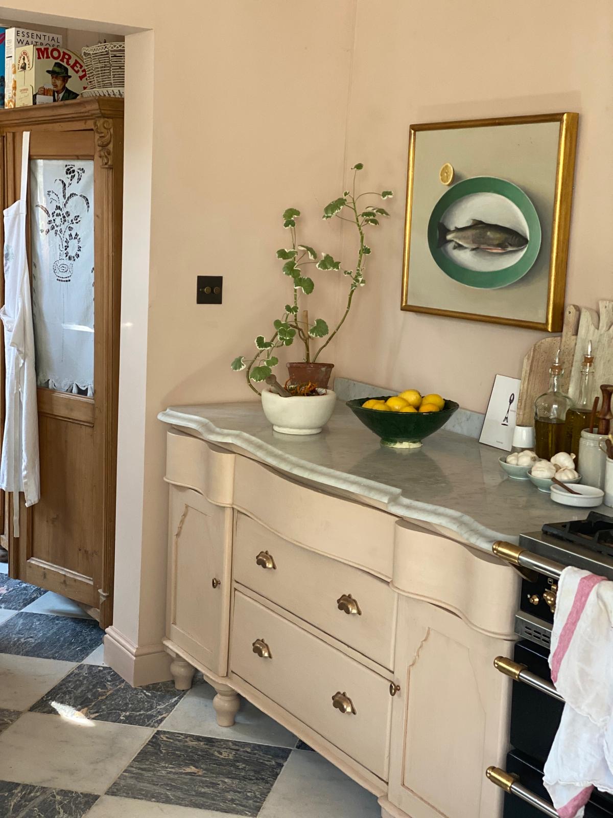 An angled photo of an ornate base cupboard, painted in a soft pink which matches the wall colour. The piece of furniture has been topped with Carrara marble. A potted plant, a ceramic bowl and some bottled oils sit on the worktop. On the wall above is a still life painting of a fish.