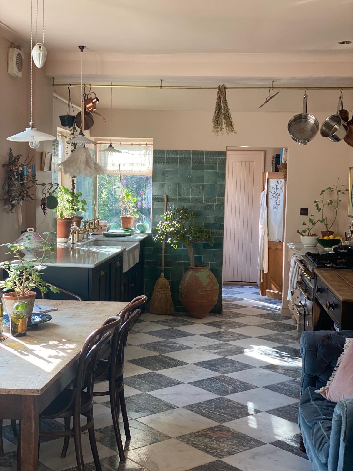 A large galley-style kitchen with cupboards along either side, as well as a dining table. The floor is made up of chequerboard tiles, the walls are a soft pink and a section of the wall straight ahead has been fitted with teal coloured tiles.