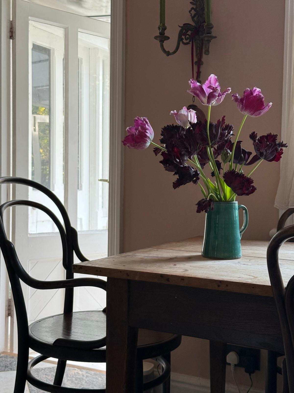 A corner of a kitchen table, it has a wooden top and a vase of pink tulips on it. The walls behind are a soft pink colour.