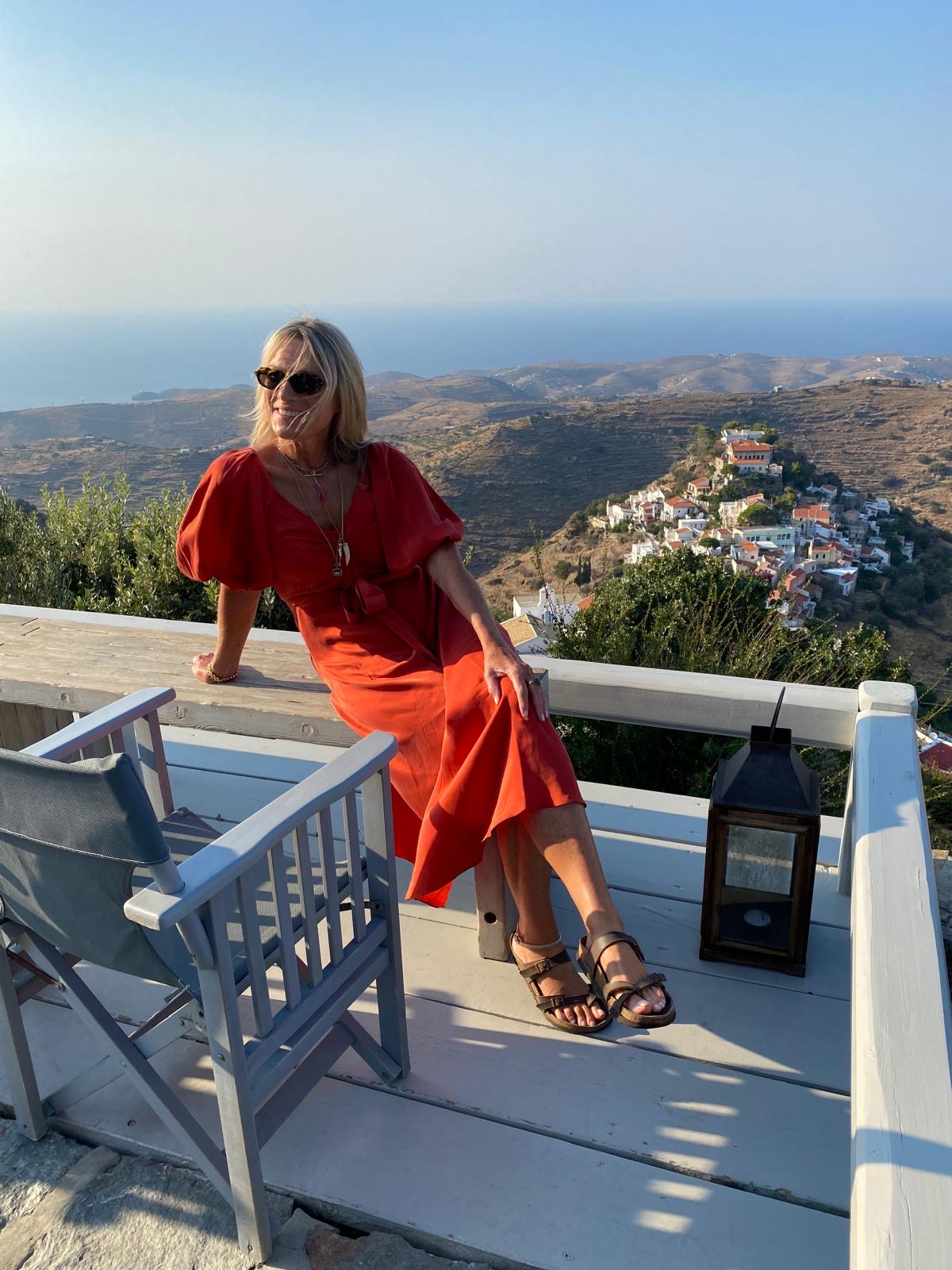 Helen Parker in a deep orange floaty dress, behind is a beautiful view of homes, hills and the sea.