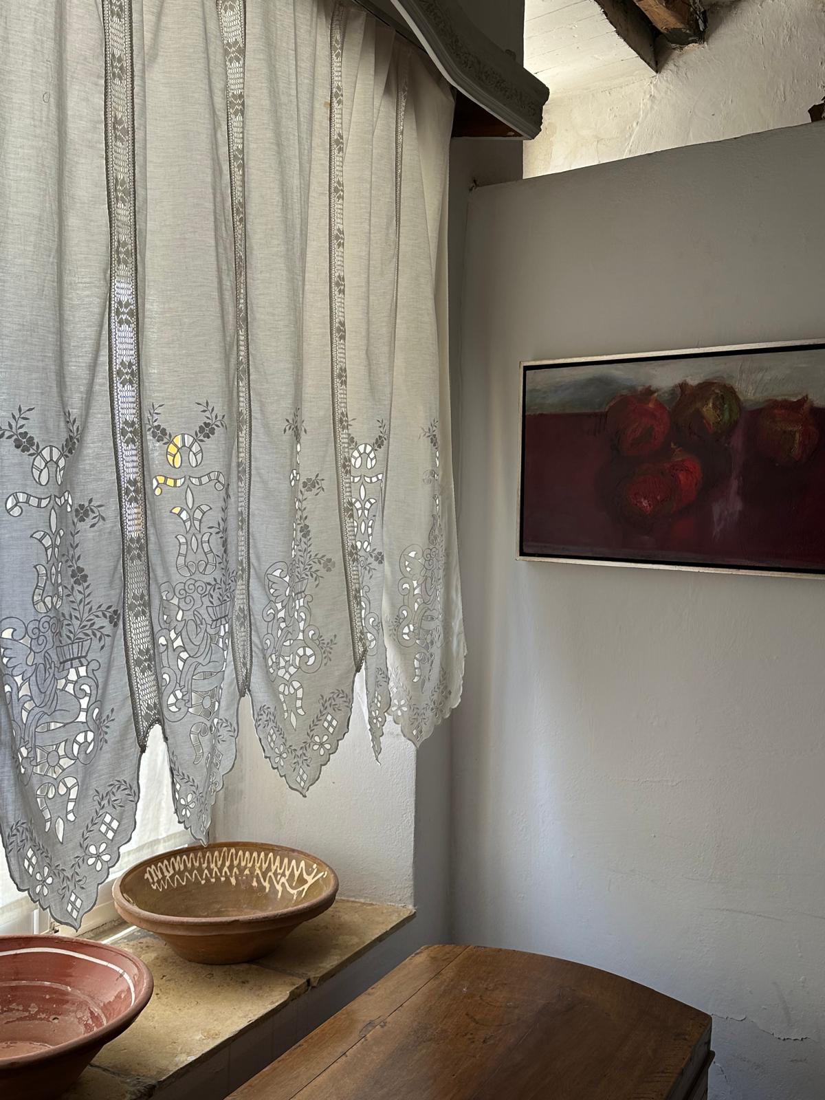 Wispy curtains hang from a window, ceramic bowls are below on the window sill. A painting has been fitted to a white wall.