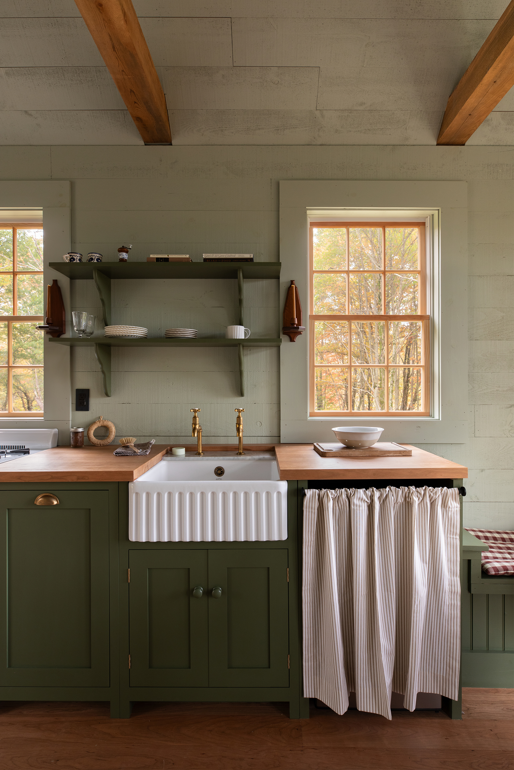 A shaker-style kitchen, it's just a short run of base cabinetry, painted in a deep green with wooden worktops. In the middle is a ceramic Belfast sink with a ribbed front. To the right is an cupboard which has a curtain front. Above are two windows and in between two shelves have been fitted. The walls have been panelled and painted in a soft green, the floorboards are bare.