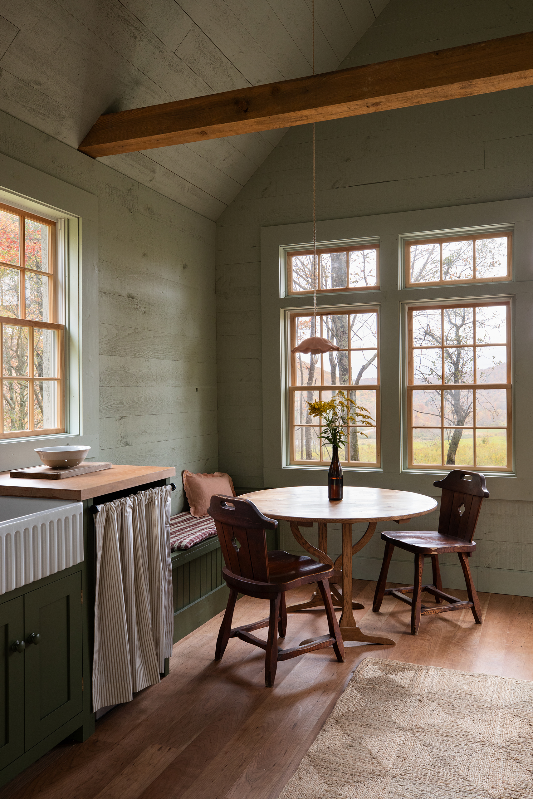 An angled photo of a little dining area in a simple space. It has a round wooden table by the window and two wooden chairs pulled up to it, against the wall a bench seat has been fitted. A small section of kitchen cabinetry is visible from the left hand side of the photo.