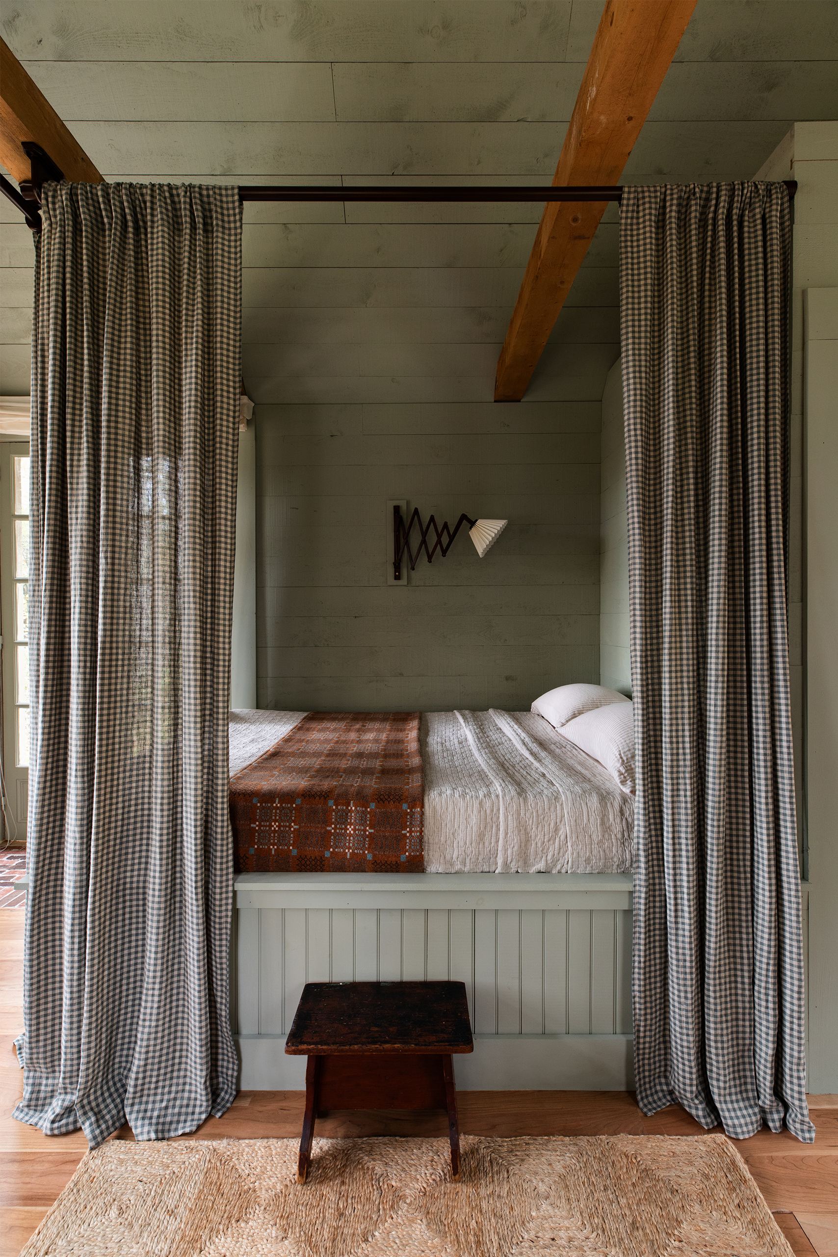 A straight on photo looking at a double bed set up. It has a panelled base and curtains fitted at the top to give it a four-poster vibe. The bedding is white and there's a brown blanket over the top. The walls and ceiling have been panelled and painted in a soft green. The floorboards have been covered with a jute rug.