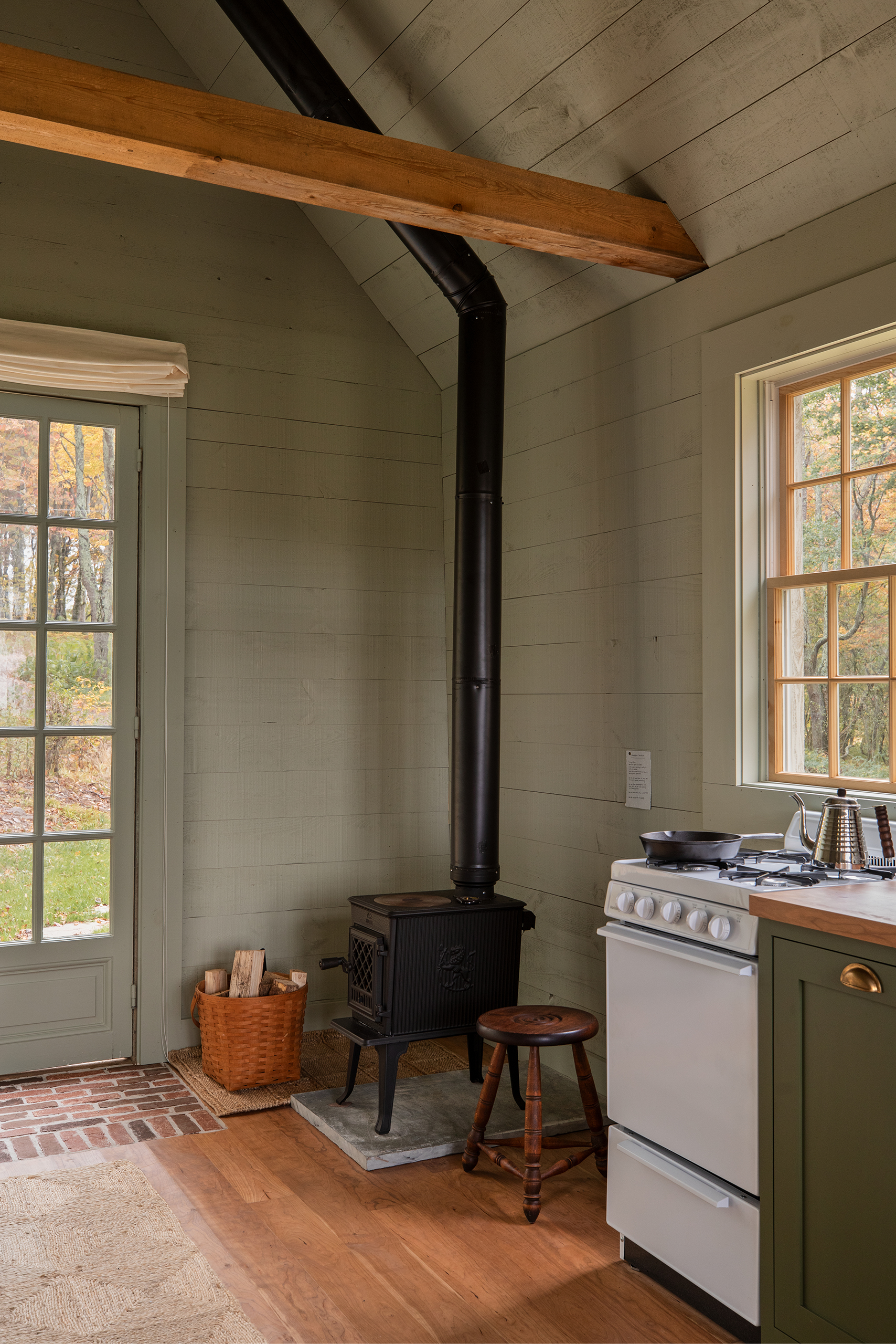 An angled photo of a wood burning stove fitted in the corner of a simple room. To its right is a white cooker. The walls have been panelled and painted in a soft green, the floorboards are bare.