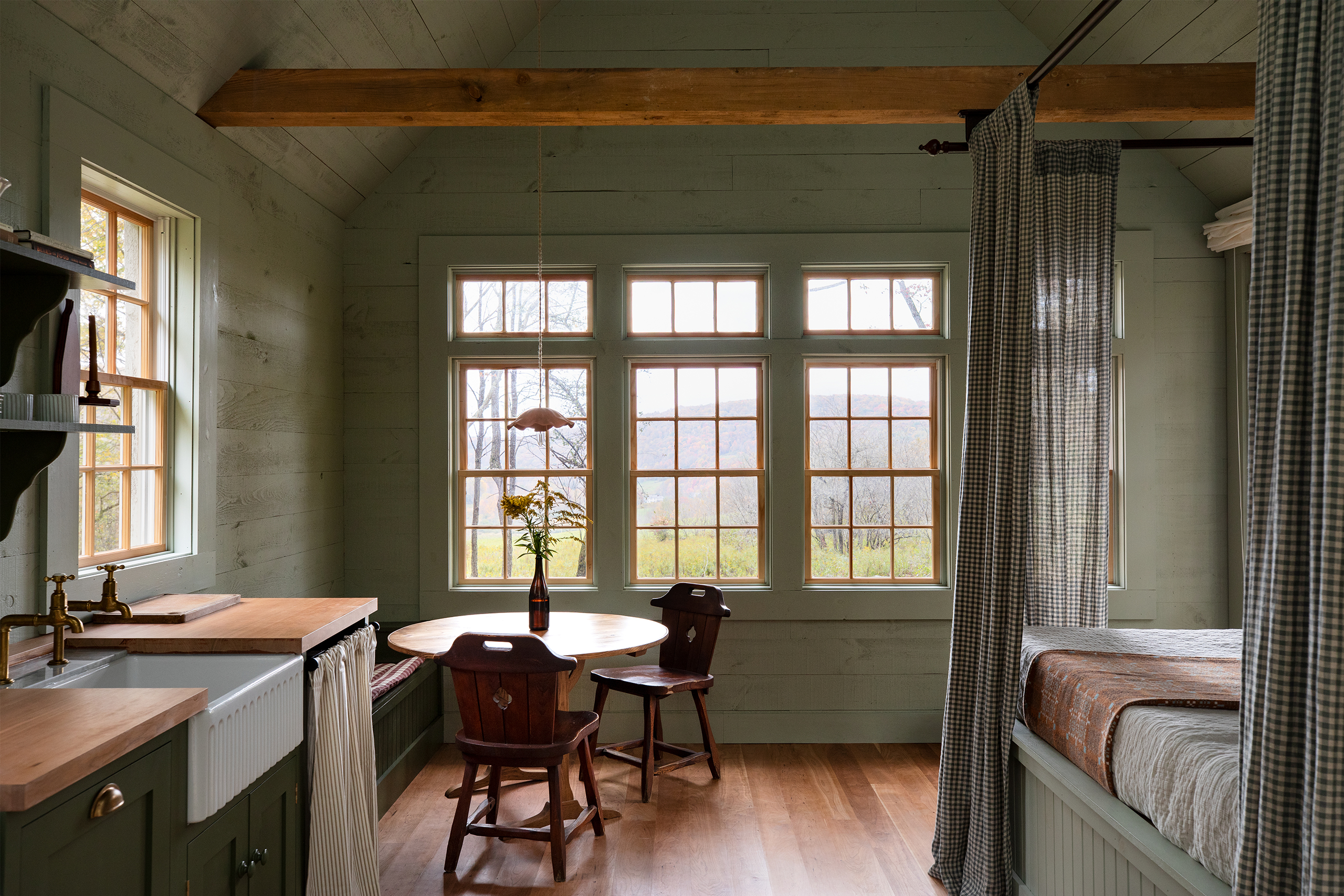 A landscape photo of a little cottage, to the left is a short run of base cabinetry, beside it is a round wooden table with seating. To the right is a bed. Straight ahead are 3 windows looking out to countryside views.