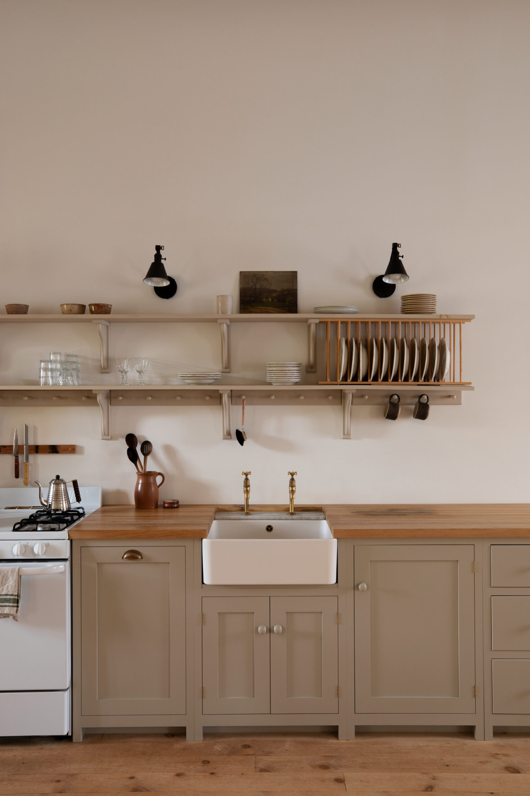 A run of shaker-style base cabinets, painted in a soft greige and topped with wooden worktops. A ceramic Belfast sink has been fitted within the run and on the far left is a white cooker. Above, two sets of open shelves have been installed, the hold kitchen aaccoutrement. The walls are an off-white colour and the floorboards are bare.