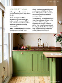 A page of a magazine, an image of a green kitchen with copper worktops, pale pink walls and a wooden floor takes up the entire page. A section of the top half of the page is taken up by a white text box with black text written inside.