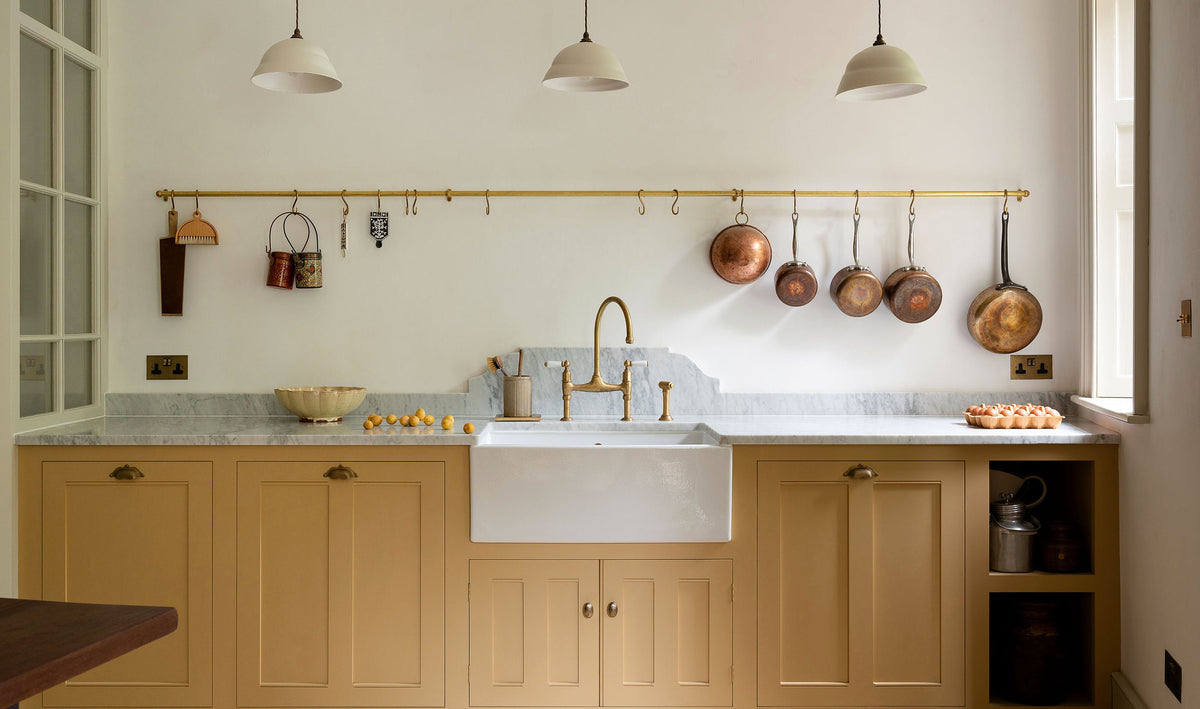 A charming blush yellow, classic English kitchen features a Carrara marble countertop, an ornate backsplash, a long aged brass rail and an aged brass tap. Decorated with copper pots, vintage ceramics, and small wooden cleaning supplies, it creates an elegant and tidy atmosphere.