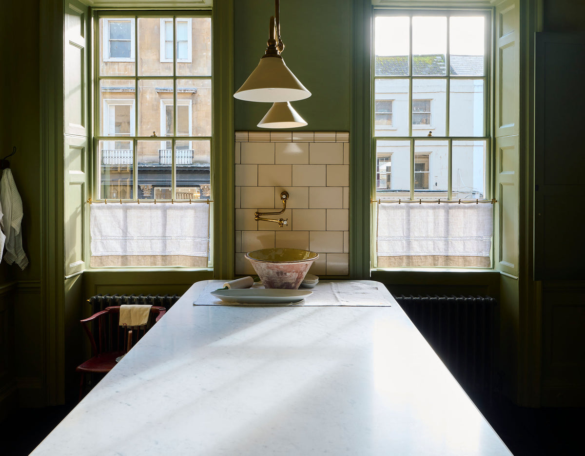 A quaint and stylish Wilkes green Heirloom kitchen features a large dairy table. Adorned with an aged brass pot filler, artisan ceramics, Heirloom gaselier lights, and a pair of aged brass curtain rail, this kitchen creates a luxurious dining and social space.