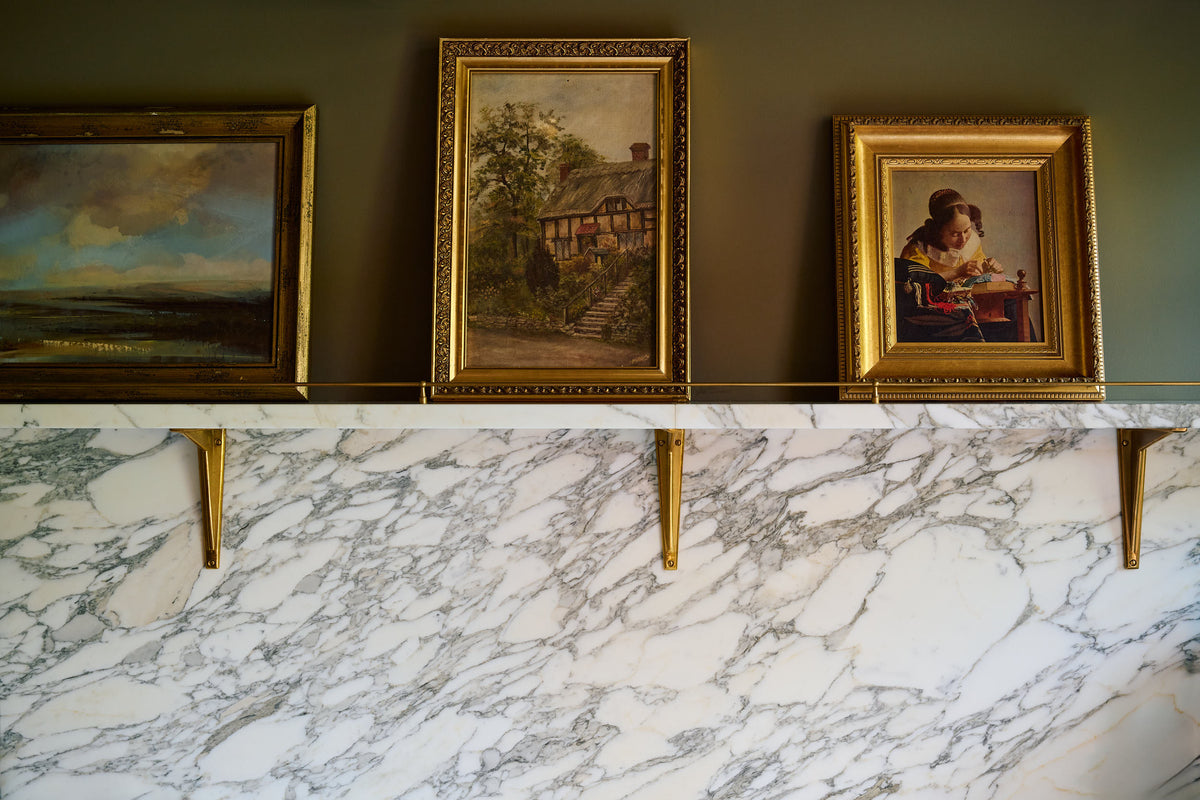 A closer look at the grand and atmospheric olive green classic English kitchen featuring a large marble splash back to shelf wall, held up with a solid brass classic shelf brackets and adorned with vintage paintings.