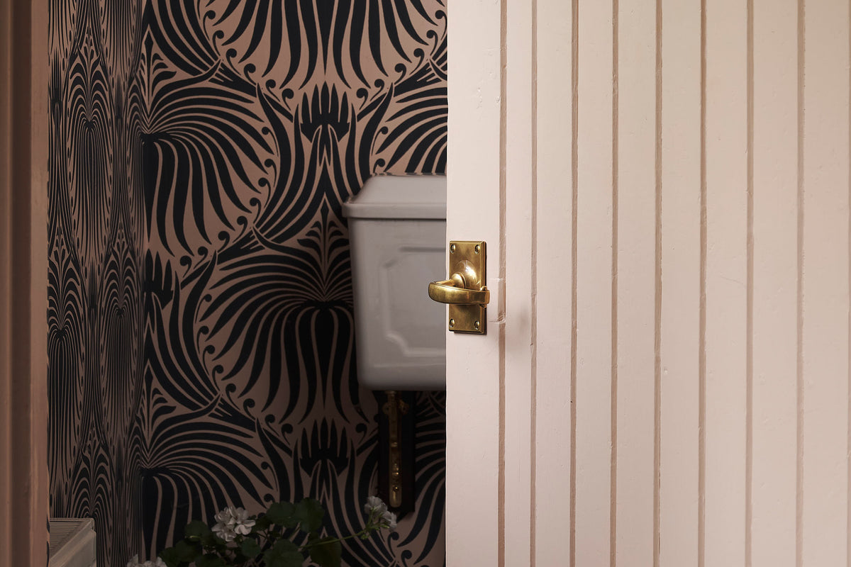 A close-up of the solid brass Otillie door handle with an Heirloom Brass finish. Promptly fixed on a Princelet pink door that is slightly opening into a small room with a pink wallpaper with black floral detail pattern.