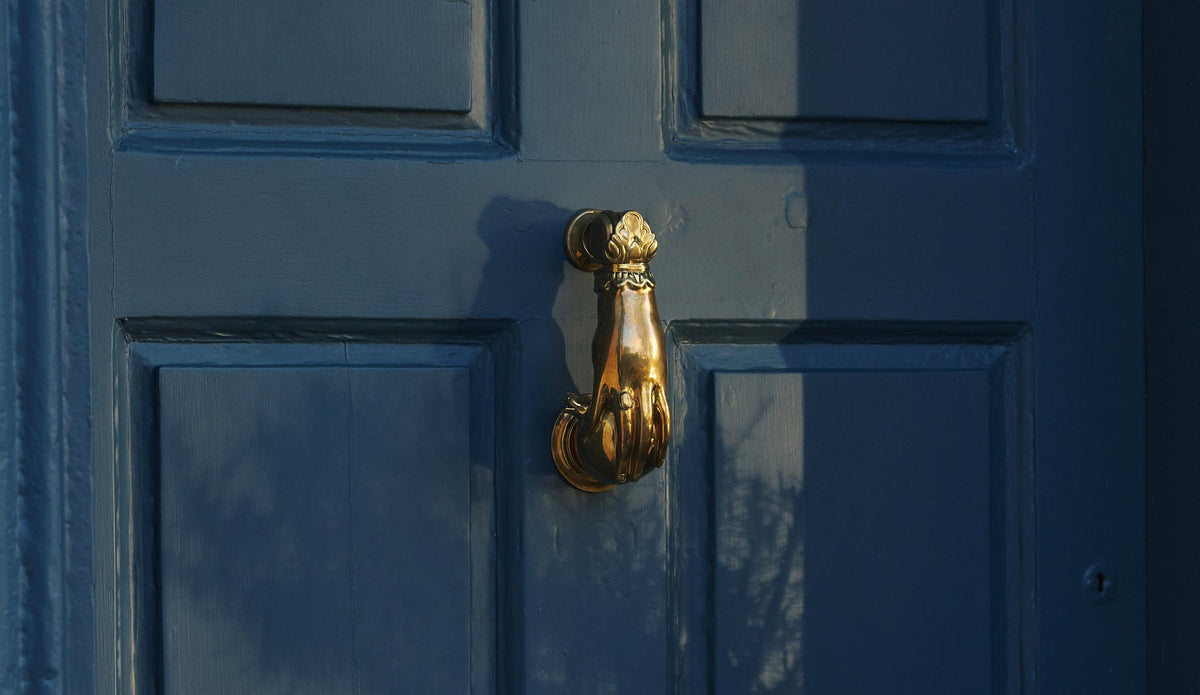 An oblique view of a decorative brass door knocker, shaped like a hand holding a pomegranate and fitted to a blue painted door.