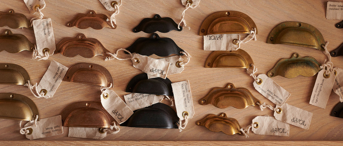 A collection of brass and bronze cabinet and drawer handles in a variety of finishes, each handle has a small canvas fabric tag labelled 'deVOL'.