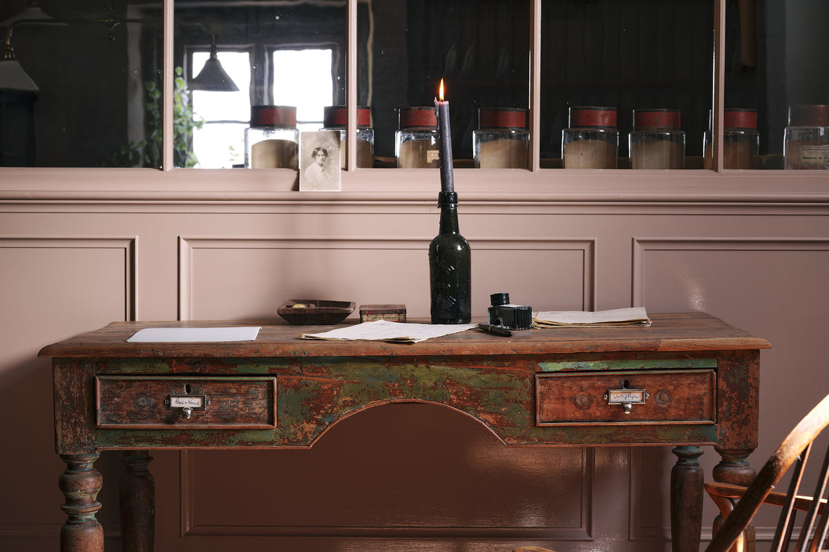 Antique silver pulls are attached to a vintage desk with chippy green paint so much of the original timber shows through. On the desk is a dark glass bottle with a lit candle, and various vintage papers, as well as an ink pen and ink bottle. The pulls themselves are a dark silver in colour, each with a paper insert describing the contents of the drawers, 'pens and pencils' and 'writing paper'.