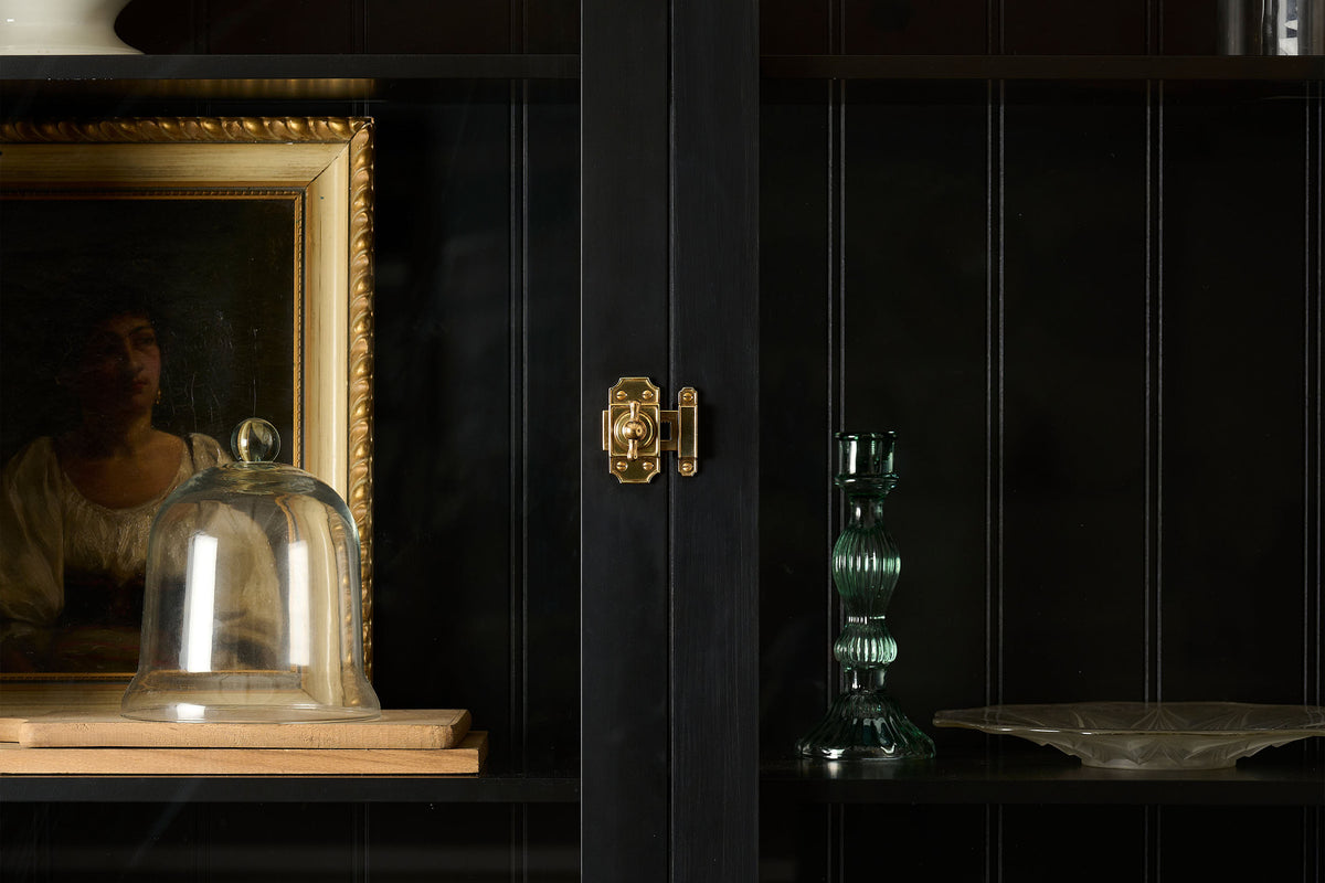 A straight-on view of a Victorian-inspired rack and pinion cupboard catch in a warm burnished brass finish, fitted to a tall glazed cupboard painted black inside and out.