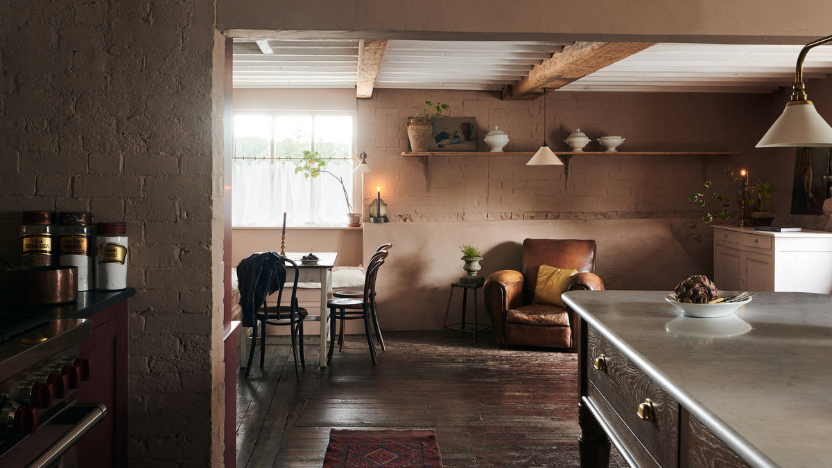 Past a refectory-red Heirloom kitchen’s cooker run and a Heirloom dairy table with an aged brass Heirloom gaselier light is a spacious dining area with vintage furniture. Beautifully decorated with a creamware pendant light, artisan ceramics, luscious green house plants, beloved candlesticks, and dried ingredients, this kitchen offers a modern twist on vintage furniture, creating a classic space designed to last.