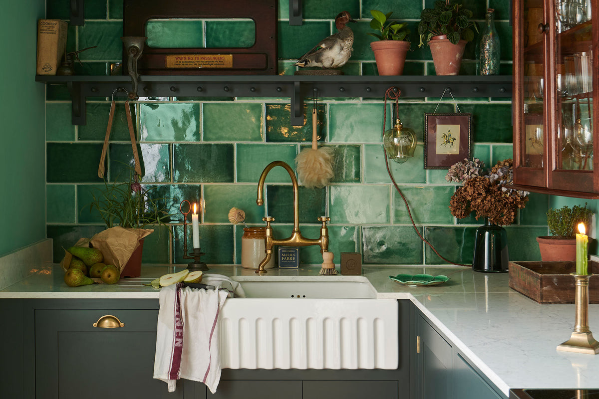 Take a closer look at the small Bakehouse Green shaker kitchen, featuring an L-shaped sink and a cooking area with a marble worktop. The vibrant Emerald Green London tiles used for the splash back add a lively touch, complemented by an array of unique decor items, including a faux taxidermy duck and a train slot window. The kitchen has a single fluted Belfast sink and an aged brass Ionian tap.
