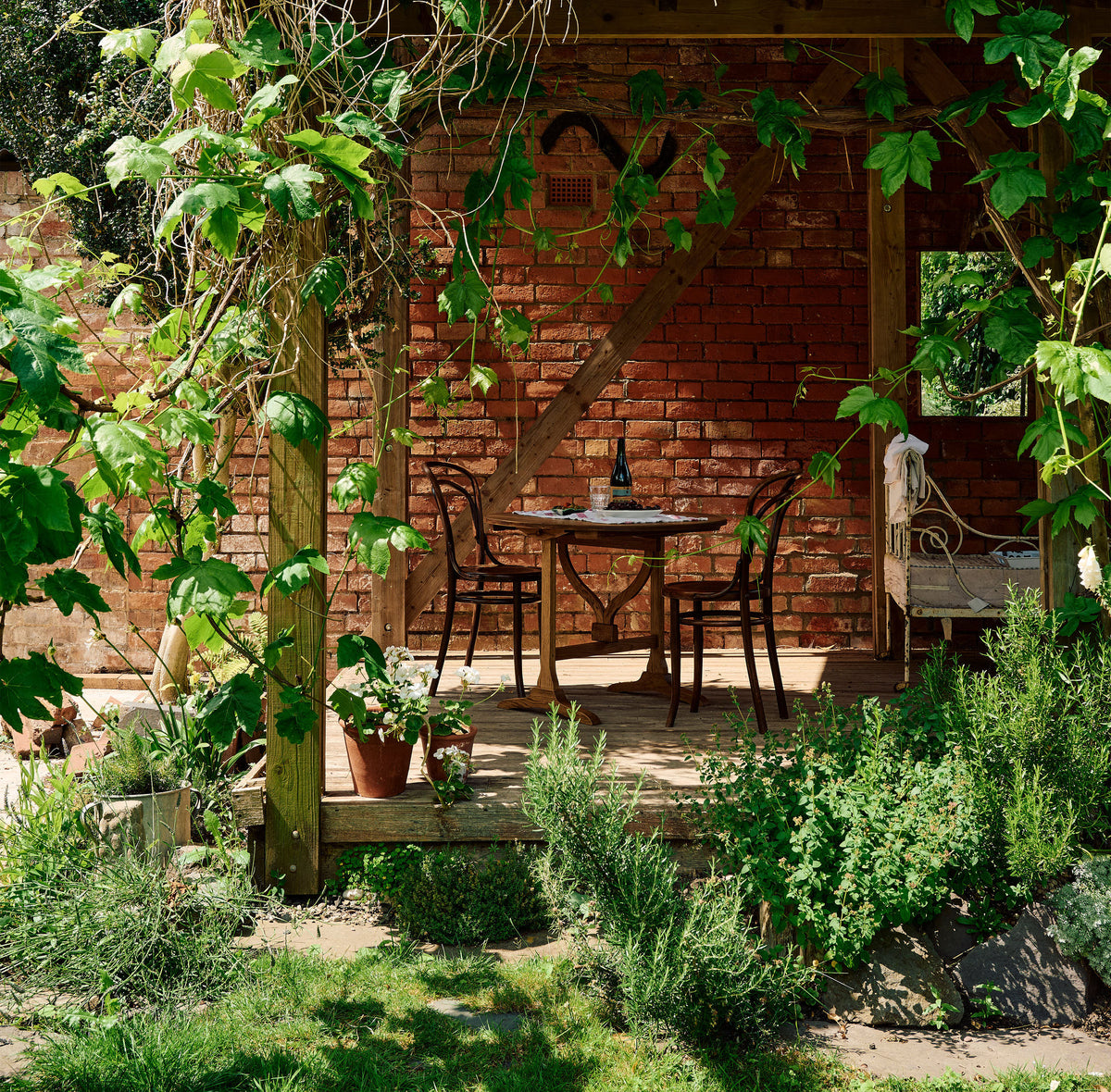 The petite vineyard table and two vintage café-style chairs outdoors on wooden decking, surrounded by lush climbing vines.