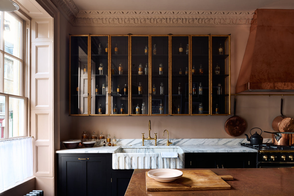 A bold and dramatic Haberdasher’s kitchen with a sink and cooker run, an Arabescato worktop and single fluted sink, an aged brass Ionian tap, a glazed wall cupboard, a large copper canopy, and a kitchen island with a copper worktop. Adorned with copper pans, artisan ceramics, labelled glass jars, and a quirky framed illustration, creating a local bristo bar in a Manhattan space.