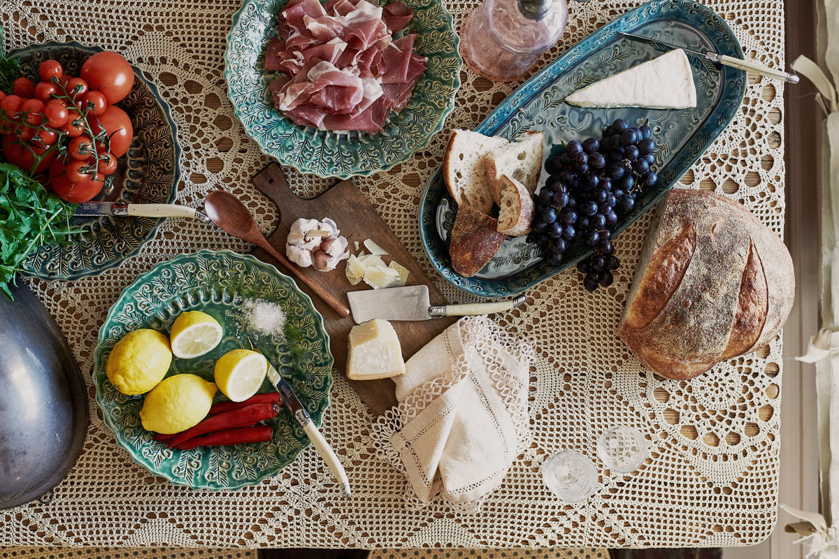 An overhead view of several oval and round handmade bohemian-style ceramic platters, with black grapes, cheeses, homemade bread, lemons, chillies and prosciutto ham on a beautiful crochet lace tablecloth.