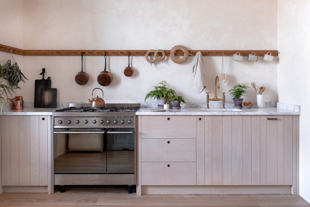 A light contemporary kitchen with creamy textured walls, rough-sawn beech slatted cupboard doors, an aged brass tap and a span of wooden pegs for hanging mugs, pans and utensils above a mottled white marble worktop. The styling is simple, Scandinavian and slightly rustic, with small potted ferns and woven rope trivets, and a built-in oven.
