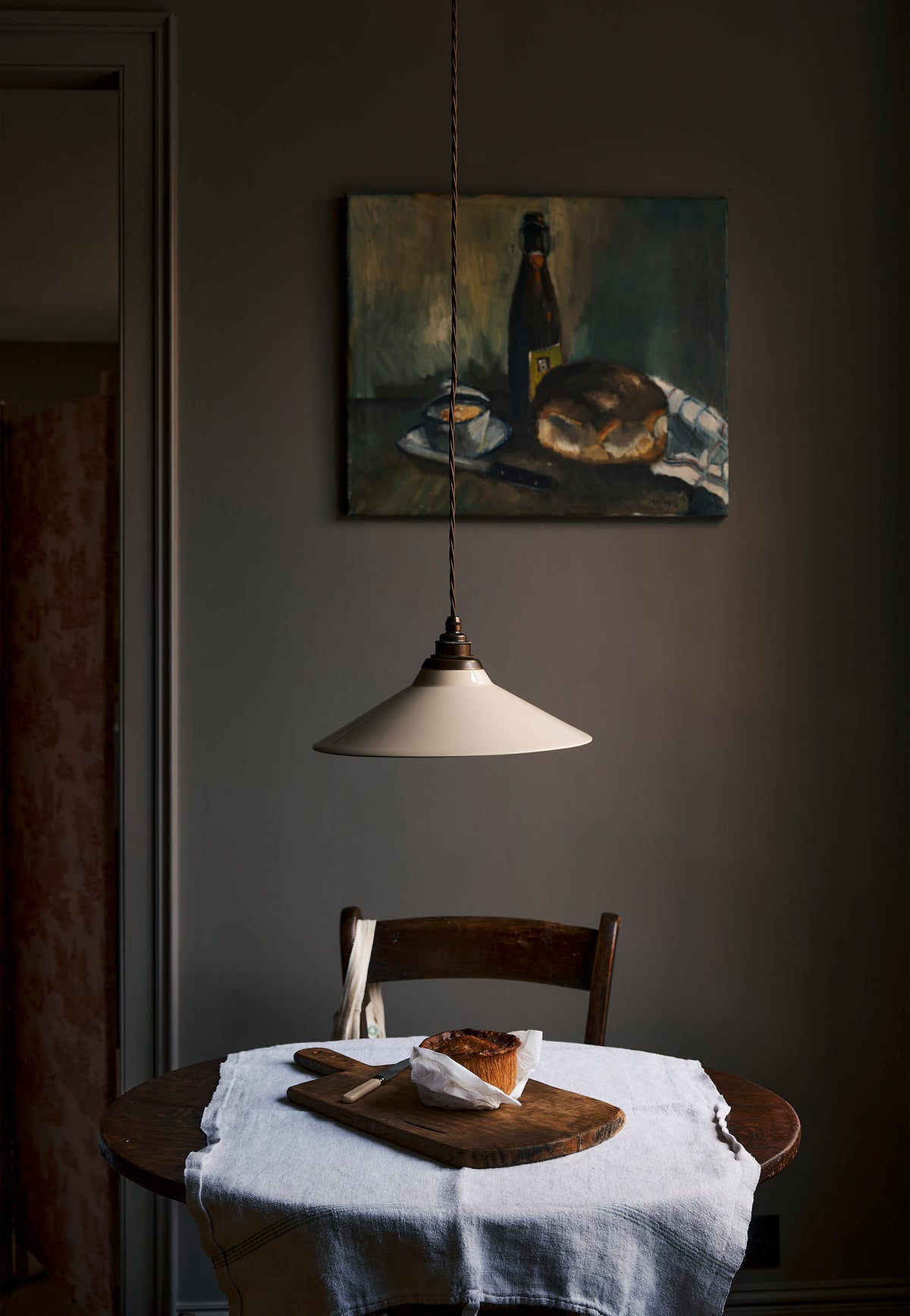 A straight-on view of a rise and fall light with a small cream glazed ceramic shade, aged brass fittings and brown twisted fabric flex cord hanging in a dark room over a small round wooden table with a linen tablecloth and fresh bread.