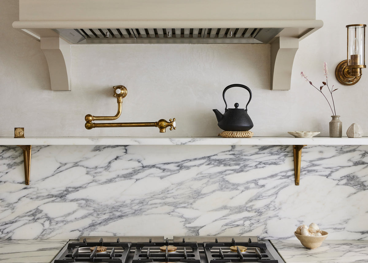 A closer look at an elegant mushroom-brown, classic English kitchen features a cooker run with an Arabescato marble worktop, splashback, and shelf. Beautifully decorated with artisan ceramics, brass classic shelf brackets, a deVOL aged brass pot filler, and a cast-iron teapot, it adds a sophisticated touch to the elegant kitchen.