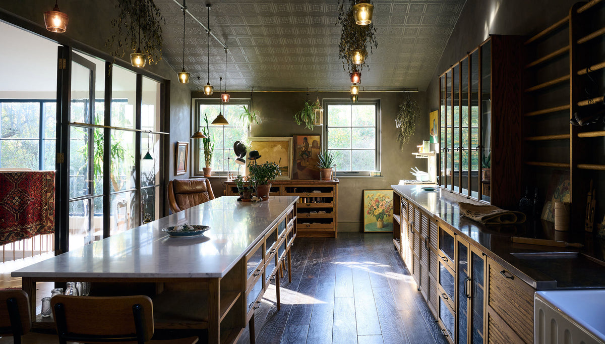 A warm and inviting Haberdasher’s kitchen features a long sink run with a Carrara marble and copper worktop, a double-fluted marble sink, a kitchen island, wall cupboards, and countertop cupboards. Adorned with luscious green hanging and potted plants, vintage items, artisan ceramics, illustrative tea towels, paintings, and a beloved white gas stove, this kitchen creates a lovely exhibitionist space that is both beautiful and practical.