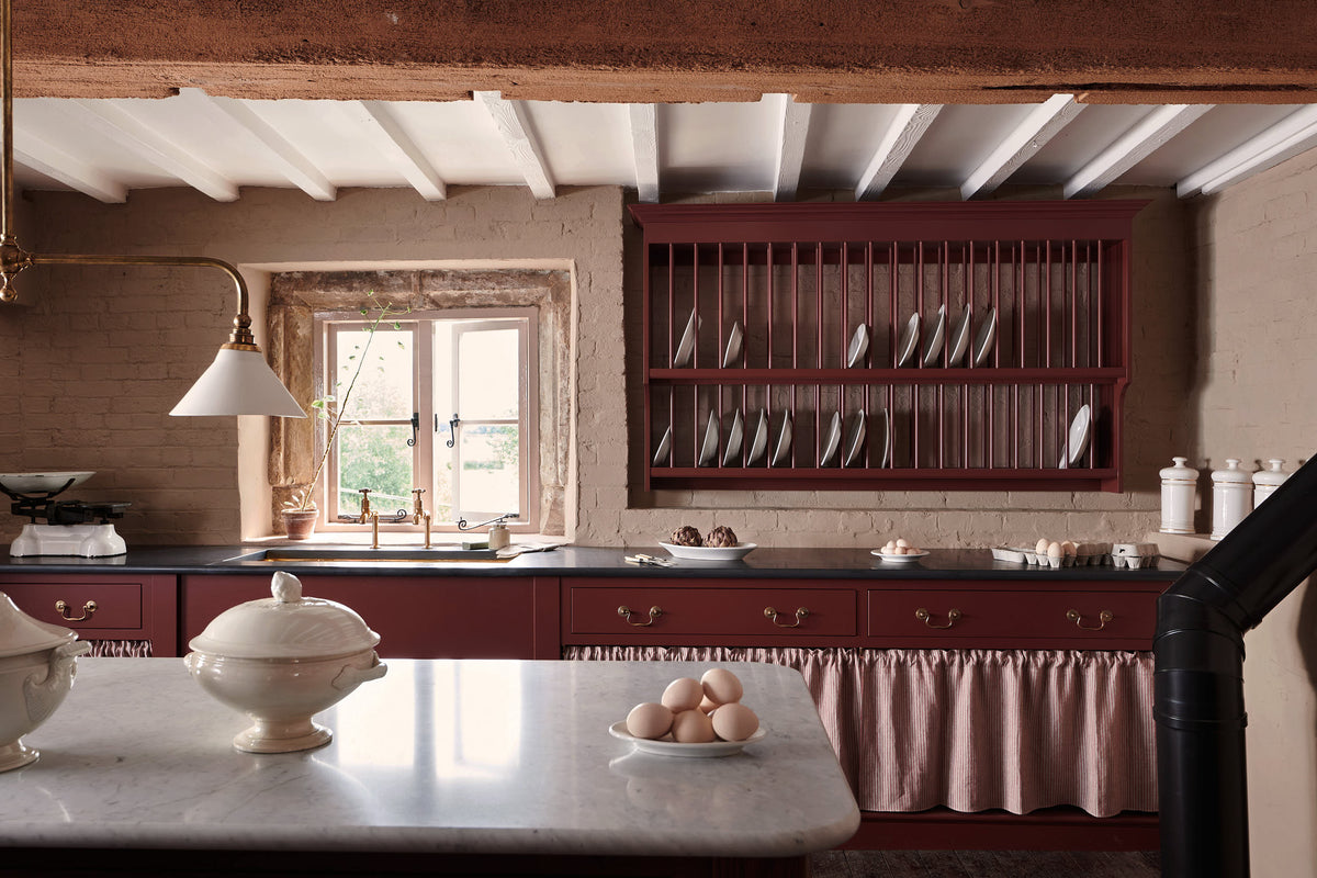 A refectory-red Heirloom kitchen’s sink cabinet reveals a polished brass sink, aged brass Mayan taps, café curtain rails, a red and white striped curtain, a plate rack, and an Heirloom dairy table. Beautifully decorated with Heirloom gaselier lights, a vintage scale, artisan ceramics, and a plate of fresh eggs, this kitchen offers a modern twist on vintage furniture, creating a classic space designed to last.