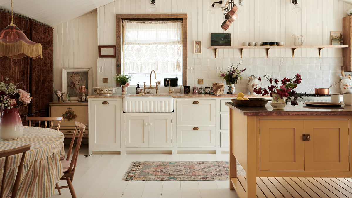 Looking straight towards the back wall of a bohemian-style kitchen that has tongue and groove panelled walls, solid floorboards, and a run of simple Shaker furniture, all painted in a creamy white shade. A mustardy yellow prep table sits in the centre of the room, creating a galley-style working area. The kitchen is fitted with soft pink marble worktops and styled with a collection of vintage pots and pans.