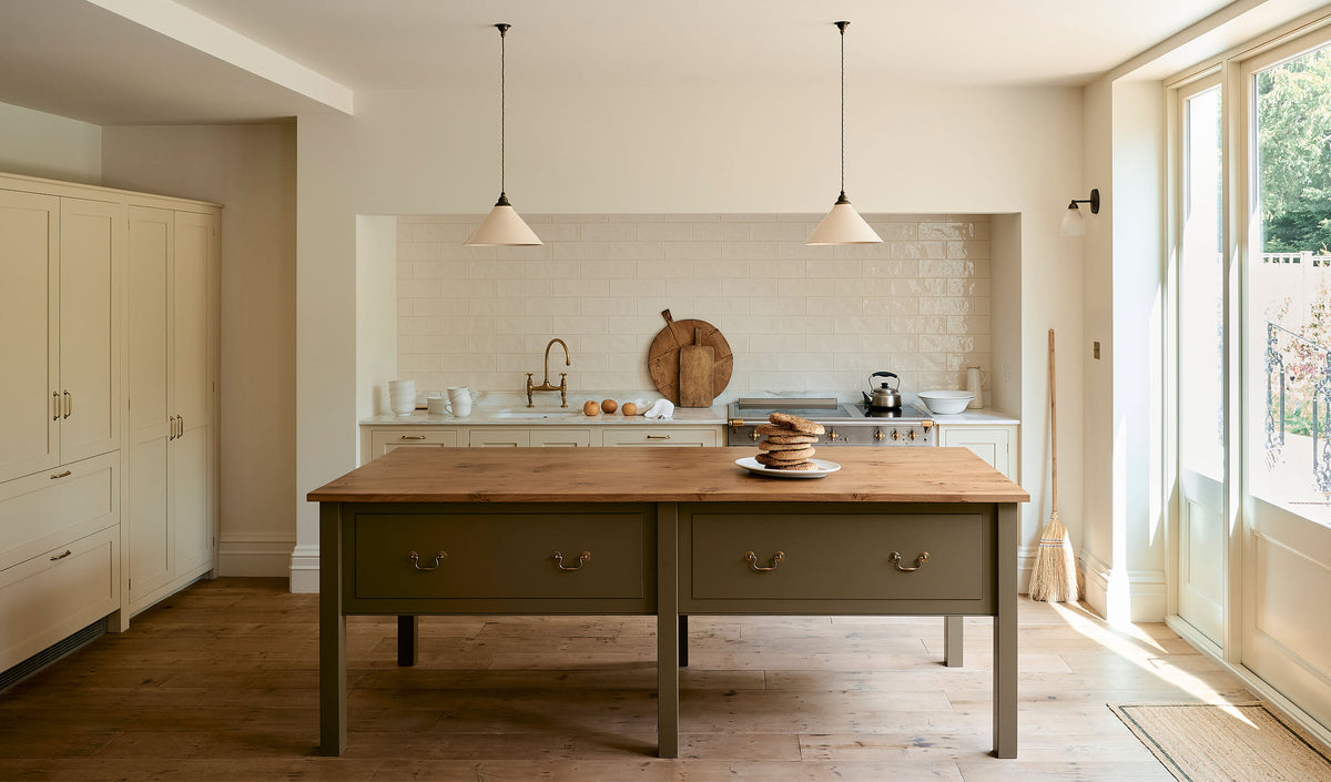 A light, clean, and simple shaker kitchen features a sink and cooker run nestled in a chimney breast, cream porcelain splashback tiles, wooden floorboards, and white walls. Cream cabinets complement an olive green Georgian-style prep table with deep drawers and a rustic oak worktop at the room’s centre, illuminated by two elegant creamware pendant lights.