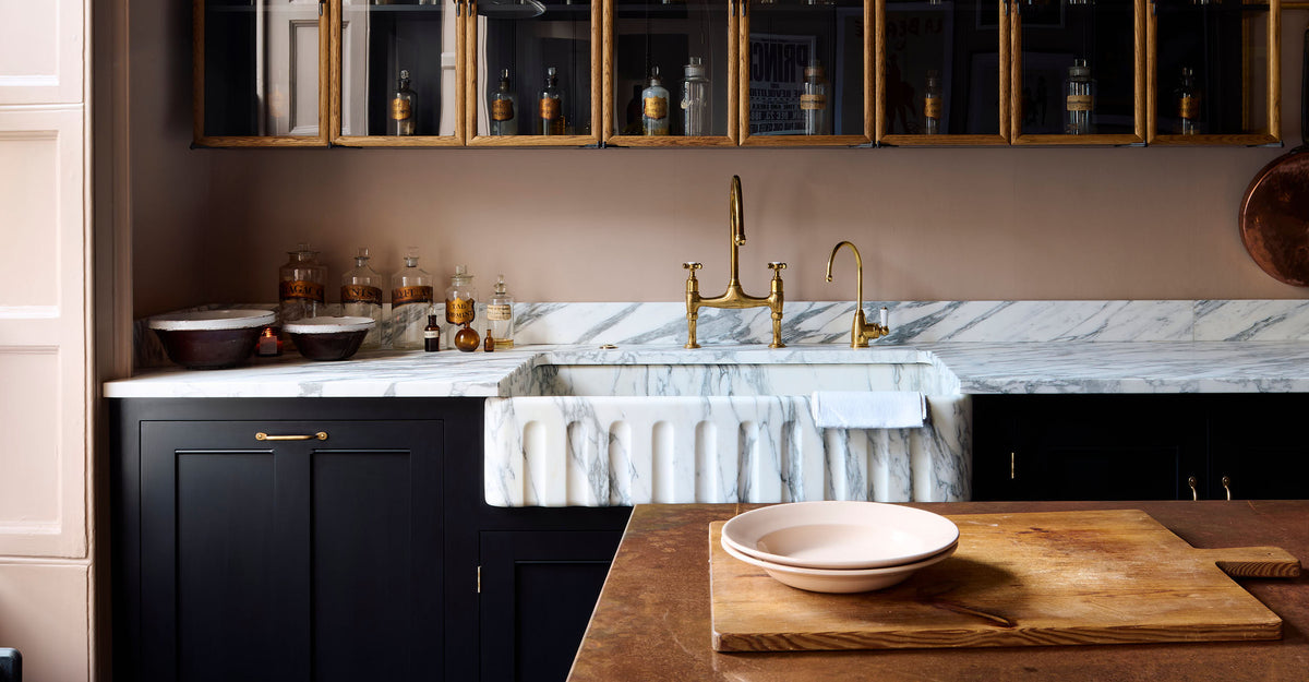 A bold and dramatic Haberdasher’s kitchen with a sink and cooker run, an Arabescato worktop and single fluted sink, an aged brass Ionian tap, a glazed wall cupboard, a large copper canopy, and a kitchen island with a copper worktop. Adorned with copper pans, artisan ceramics, labelled glass jars, and a quirky framed illustration, creating a local bristo bar in a Manhattan space.