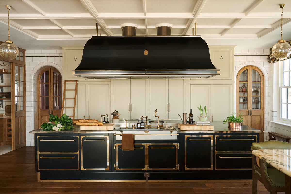 A grand room with high panelled ceiling, traditional glossy white tiled walls with curved archways on either side of a huge bank of creamy mushroom fitted full-height Classic cupboards with a polished oak ladder. An elaborate black with gold accent kitchen island and canopy sits in the middle of the space with an array of articulated brass sink taps around a wide and shallow sink.