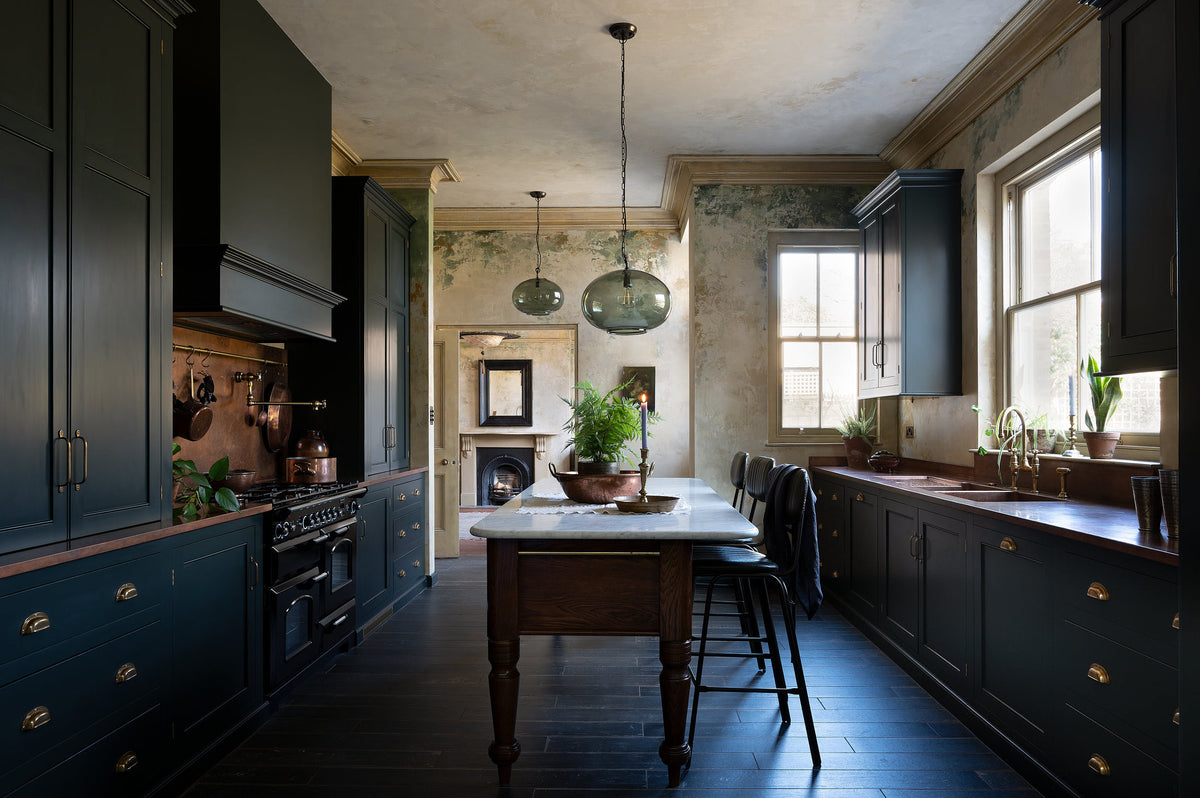 An elegant classic kitchen includes a cooker and sink with an aged copper worktop, a shaker canopy, counter cabinets, a marble worktop heirloom dairy table, a breakfast nook, and tall black chairs. The tall-ceilinged room has a rustic wash finish, with green draping down the walls, resembling overgrown ancient ruins. Beyond the archways leading to the living room is a striking black fireplace, a black-framed mirror, and a woven chandelier.