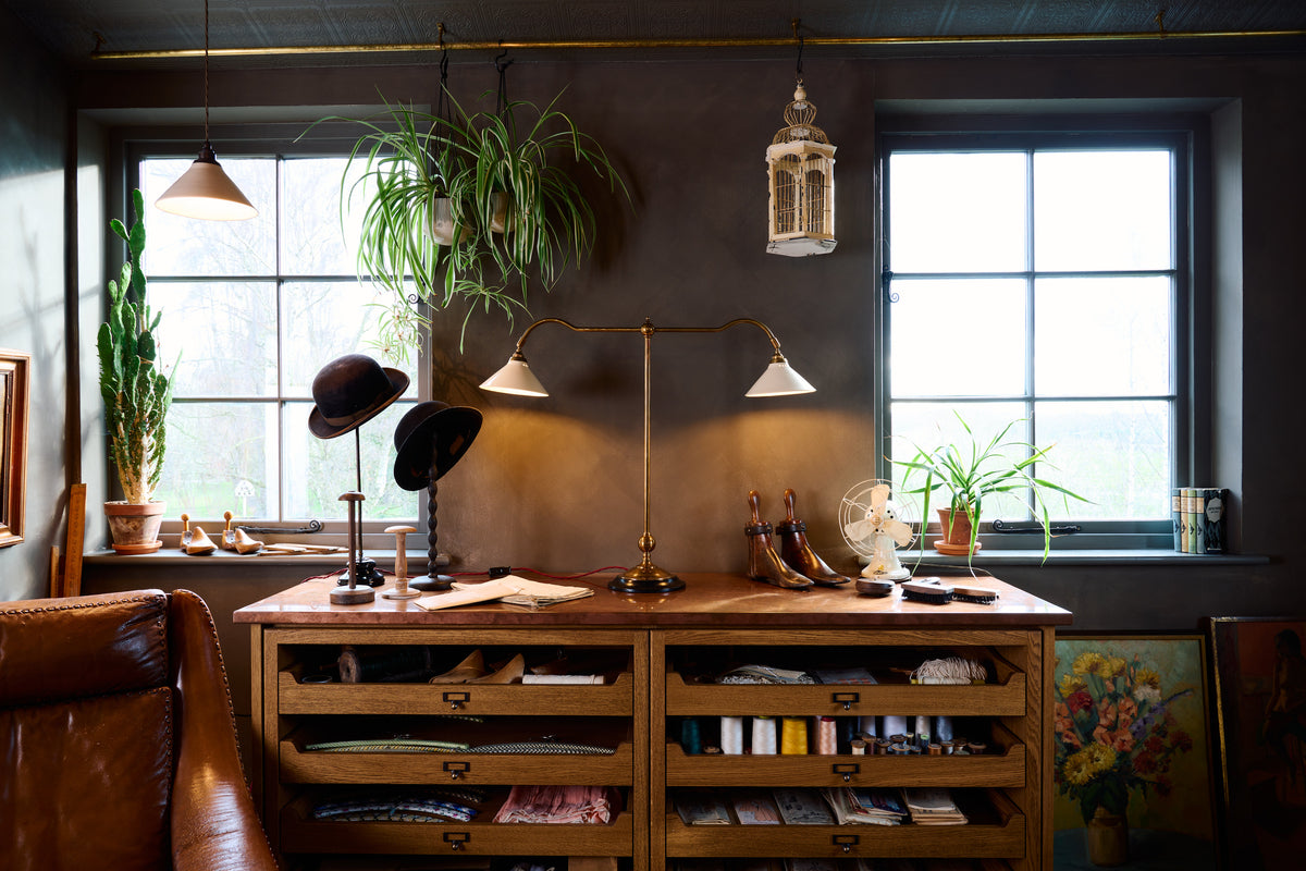 A straight-on view of a Victorian-inspired desk lamp with a solid brass foot, stand, two curved arms and two small cream glazed ceramic light shades on top of haberdasher's cupboards.