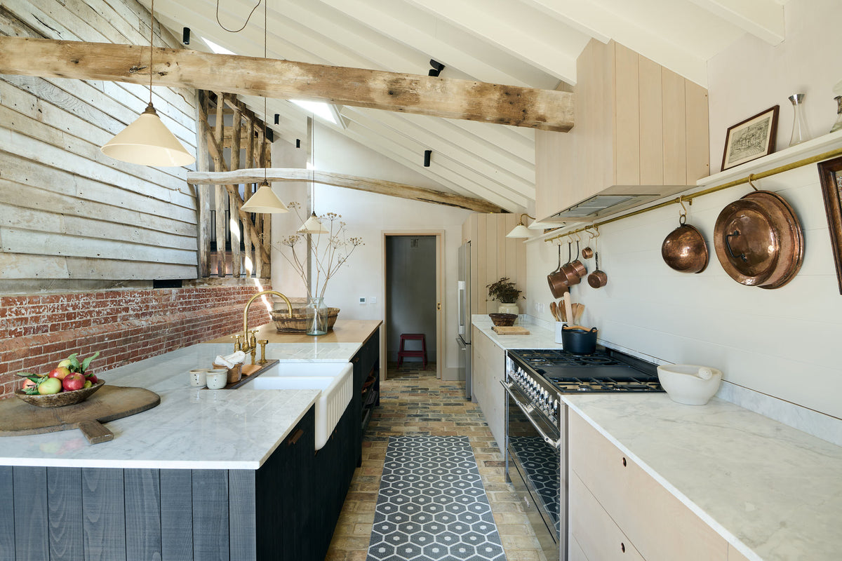 A beautifully renovated farm features a Sebastian Cox kitchen with a long cooker run, a large kitchen island with a double fluted Belfast sink, an aged brass Ionian tap, and a Carrara marble and solid oak worktop. Adorned with creamware pendant lights, vintage items, artisan ceramics, and a bowl of apples, this kitchen creates a clean and contemporary style that harmonises with the natural materials used throughout the space.