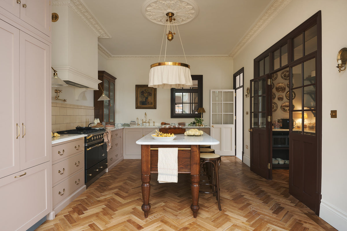 A soft Princelet pink Shaker kitchen unfolds with an L-shaped cooker and sink run topped in Carrara marble. It features a counter cupboard, full-height cabinets, cream subway tiles, a large Dairy Table surrounded by four Bentwood café stools, and glazed doors leading to a spacious pantry. Gold accents shine in the pale oak herringbone floors, aged brass taps, and a grand chandelier draped with a loose fabric shade.